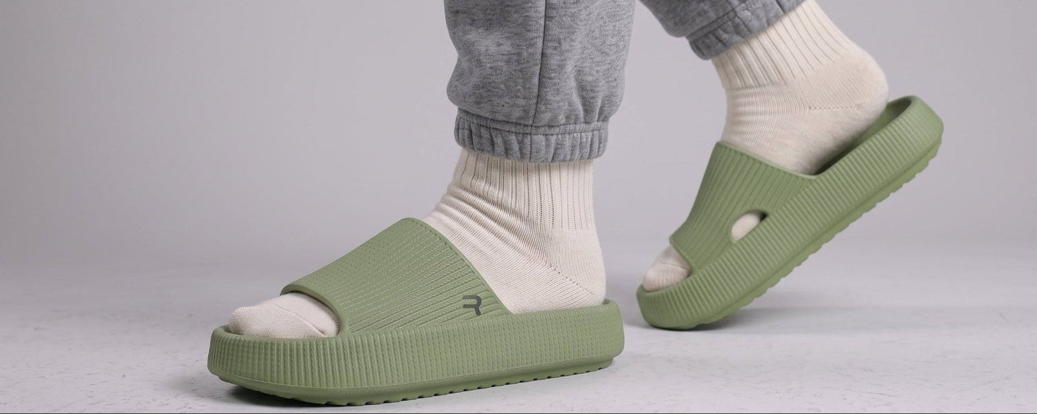 A person wearing light sage green slide sandals with white socks and gray sweatpants, shown from the calves down against a light gray studio background.