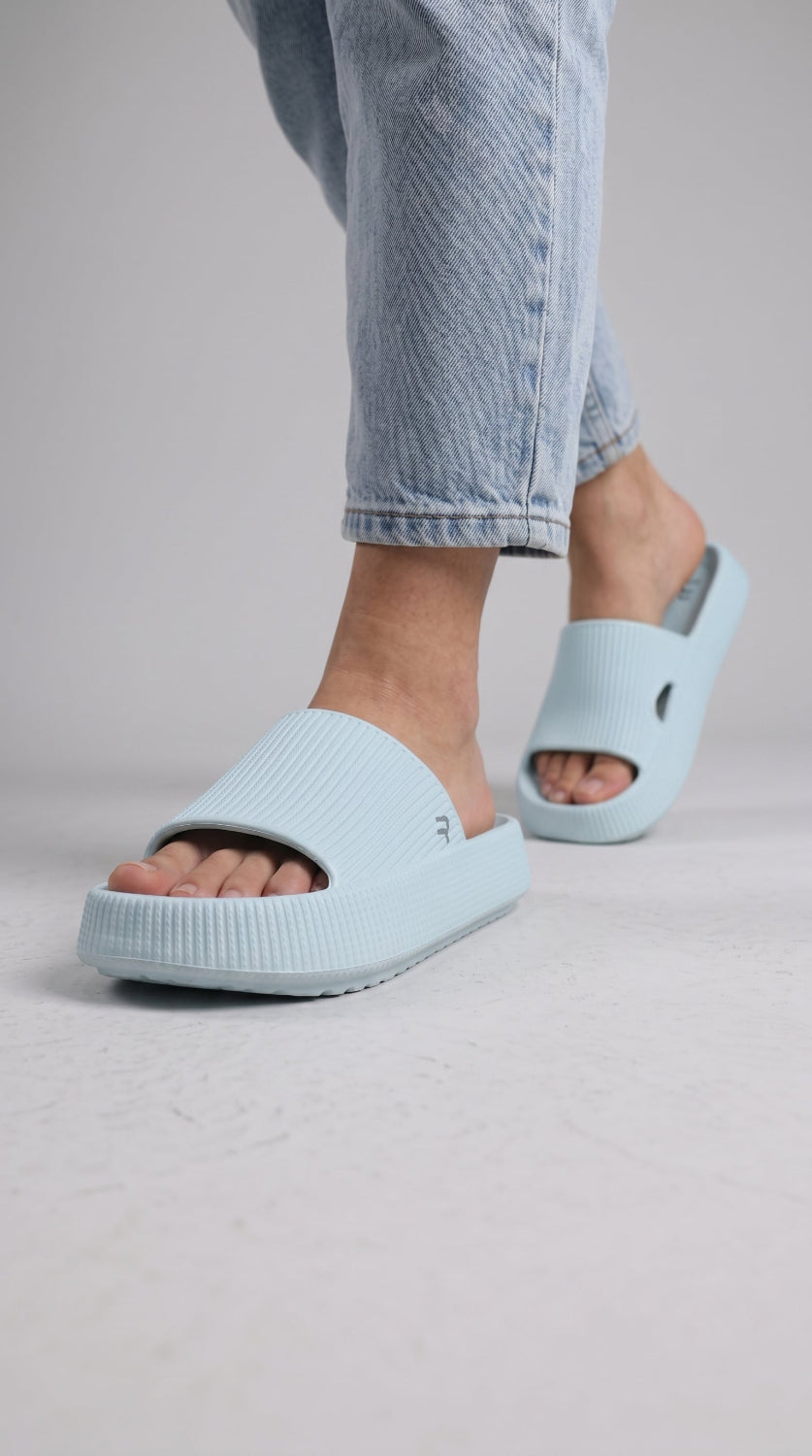 A person wearing light blue foam slide sandals with light blue jeans, shown from the calves down against a plain light gray studio background.
