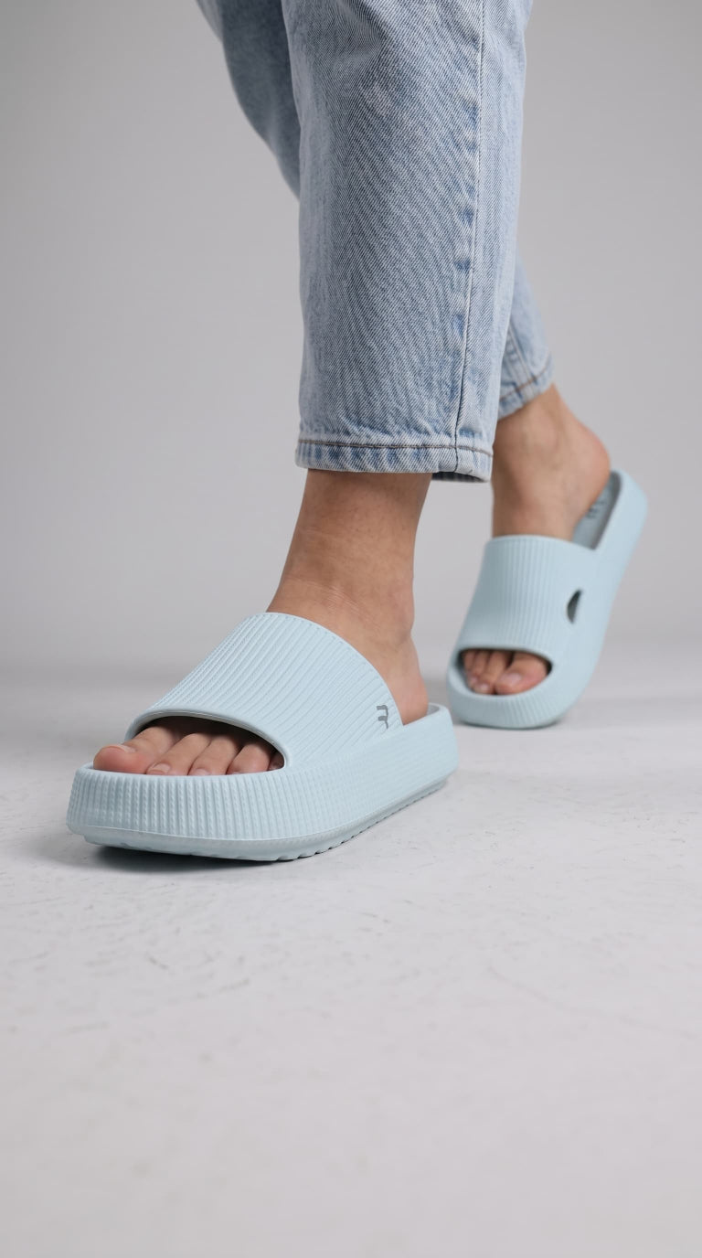 A person wearing light blue foam slide sandals with light blue jeans, shown from the calves down against a plain light gray studio background.