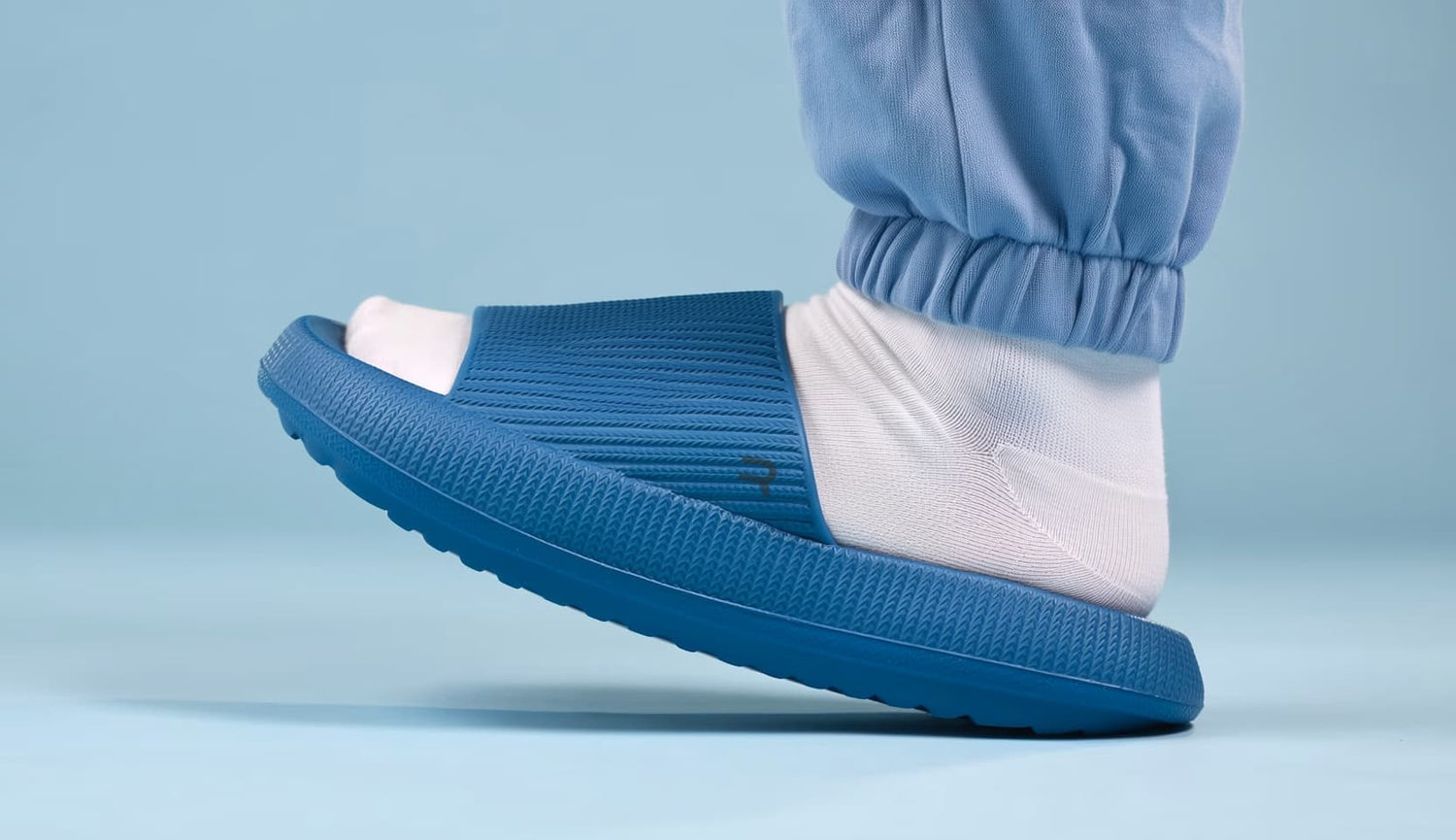 A blue slide sandal with a thick textured sole, shown worn on a foot with a white sock and light blue pants against a light blue background.