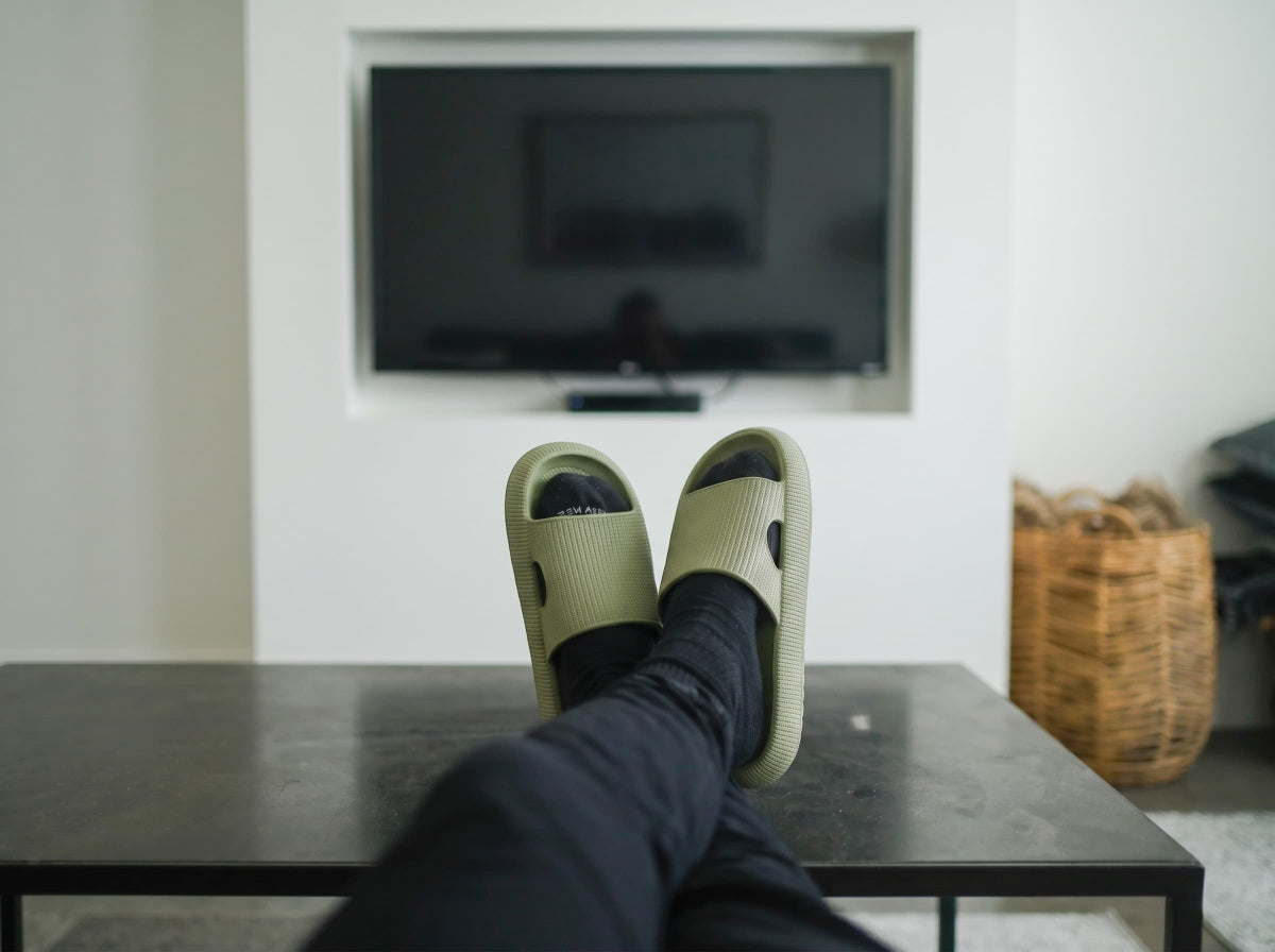 A person's feet in black socks wearing light olive green slide sandals resting on a dark table, with a blurred TV and a wicker basket in the background.