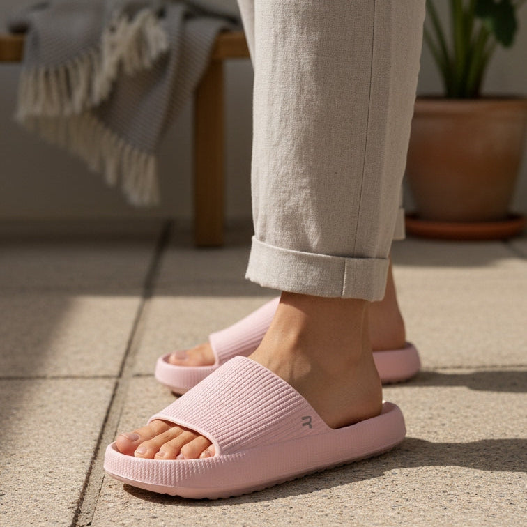 A person wearing light pink slide sandals with matching pink socks, standing on a light stone tiled floor in a sunlit indoor setting with a plant and bench in the background.